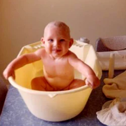 Baby Paul Hamon sitting naked in a yellow plastic bucket on a kitchen counter, laughing.