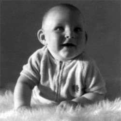 Black-and-white photo of baby Paul Hamon crawling on a fluffy rug, wearing a knitted zip-up jumper, 1973.