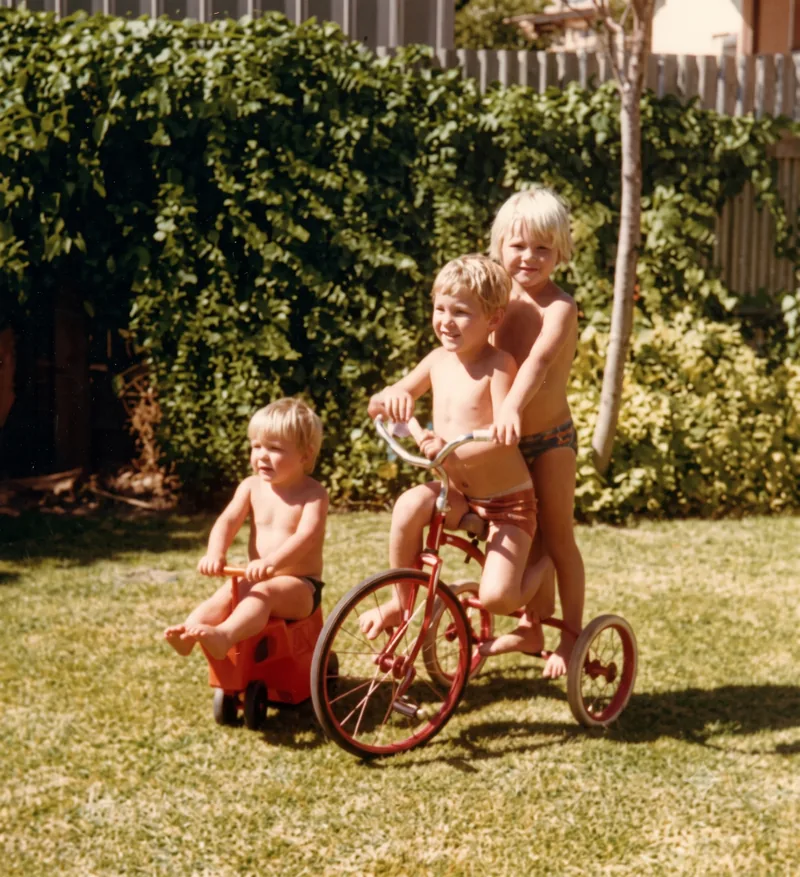 Three young children in a sunny backyard; a small child on a red ride-on and two older children on a red tricycle in front of a green hedge, Pooraka, Adelaide, summer 1977. Digitally restored version.