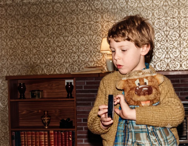 Young boy in a knitted jumper holds a soft toy bear and a small object in a lounge room with patterned wallpaper, Croydon Park, Adelaide, 1977. Digitally colourised and restored version.