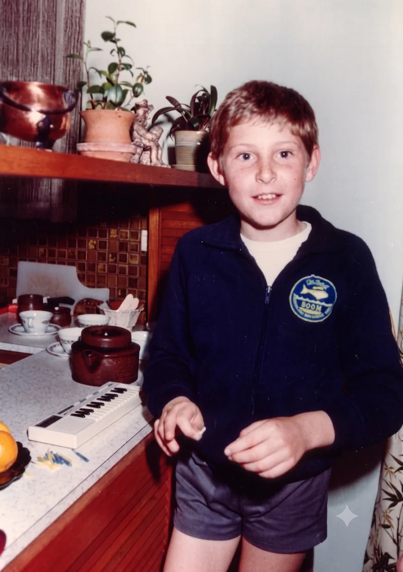 Seven-year-old Paul Hamon in a navy zip-up jacket with a swimming patch and grey shorts stands in the family kitchen, Pooraka, Adelaide, early 1980. A brown ceramic teapot and small toy piano sit on the bench. Digitally restored version.
