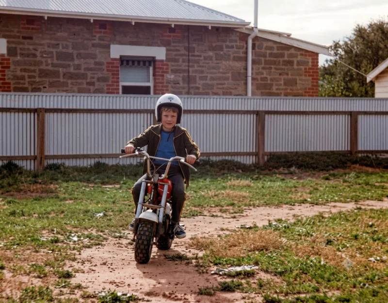 Seven-year-old Paul Hamon in a white helmet rides a small minibike on a dirt track in front of a stone house and corrugated fence, Pooraka, Adelaide, c. January 1980. Digitally restored version.