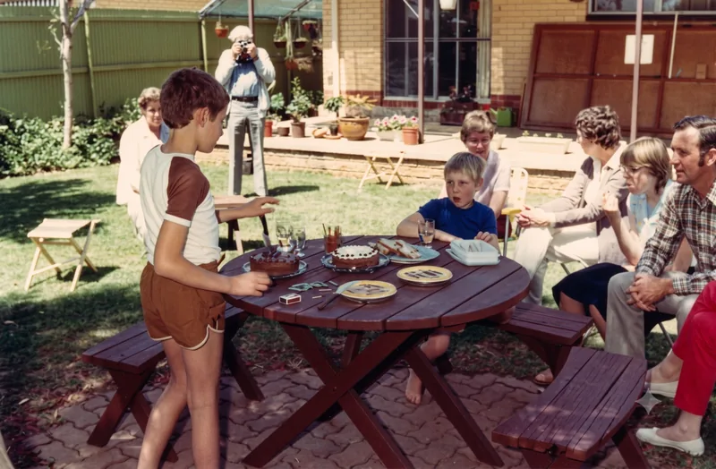 Boy in brown-and-white shirt stands at a round outdoor table with birthday cake while family members sit and stand in a backyard, Pooraka, Adelaide, November 1980. Digitally restored version (one adult male figure on the right appears AI-inserted).