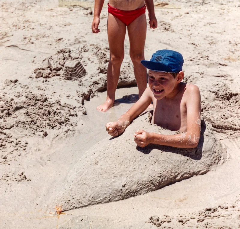 Boy buried up to his chest in a sand mound at the beach, another child’s legs visible behind, Semaphore Beach, Adelaide, summer 1980. Digitally restored version.