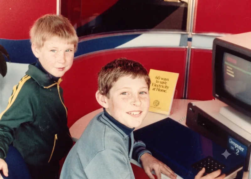 Ten-year-old Paul Hamon sits smiling at a Computer 2 terminal while seven-year-old Mark Hamon stands behind him at an ETSA display, Adelaide, c. August 1982 (possibly Royal Adelaide Show). A yellow electricity-saving booklet is on the desk. Digitally restored version.