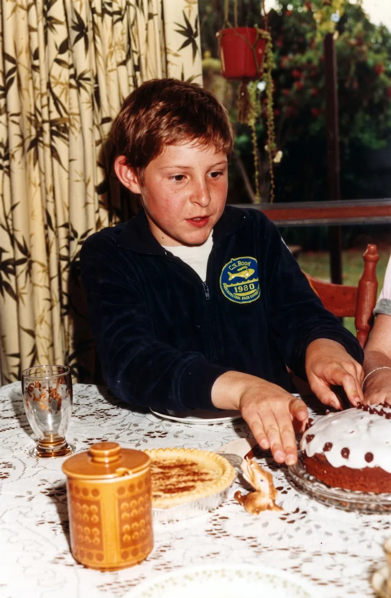 Boy in a blue jacket with swimming patches sits at a dining table reaching for a cake, Pooraka, Adelaide, November 1983. Digitally restored version.