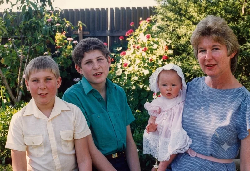 Two boys, a baby in a pink dress and bonnet, and a woman stand in a garden with roses and a wooden fence, Croydon Park, Adelaide, 1985. Digitally restored version.