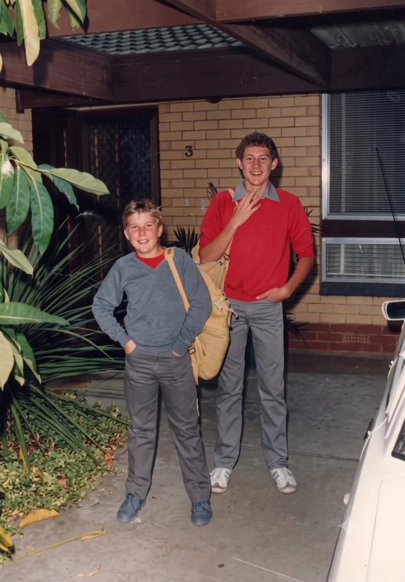 Two boys with school bags standing in front of a brick house, Pooraka, Adelaide, late January 1988. Digitally restored version.
