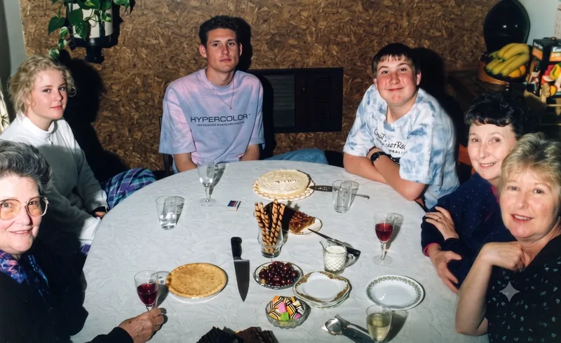 Group of six people seated around a kitchen table with cake and drinks, Pooraka, Adelaide, November 1989. Digitally restored version (Paul’s appearance enhanced by AI).